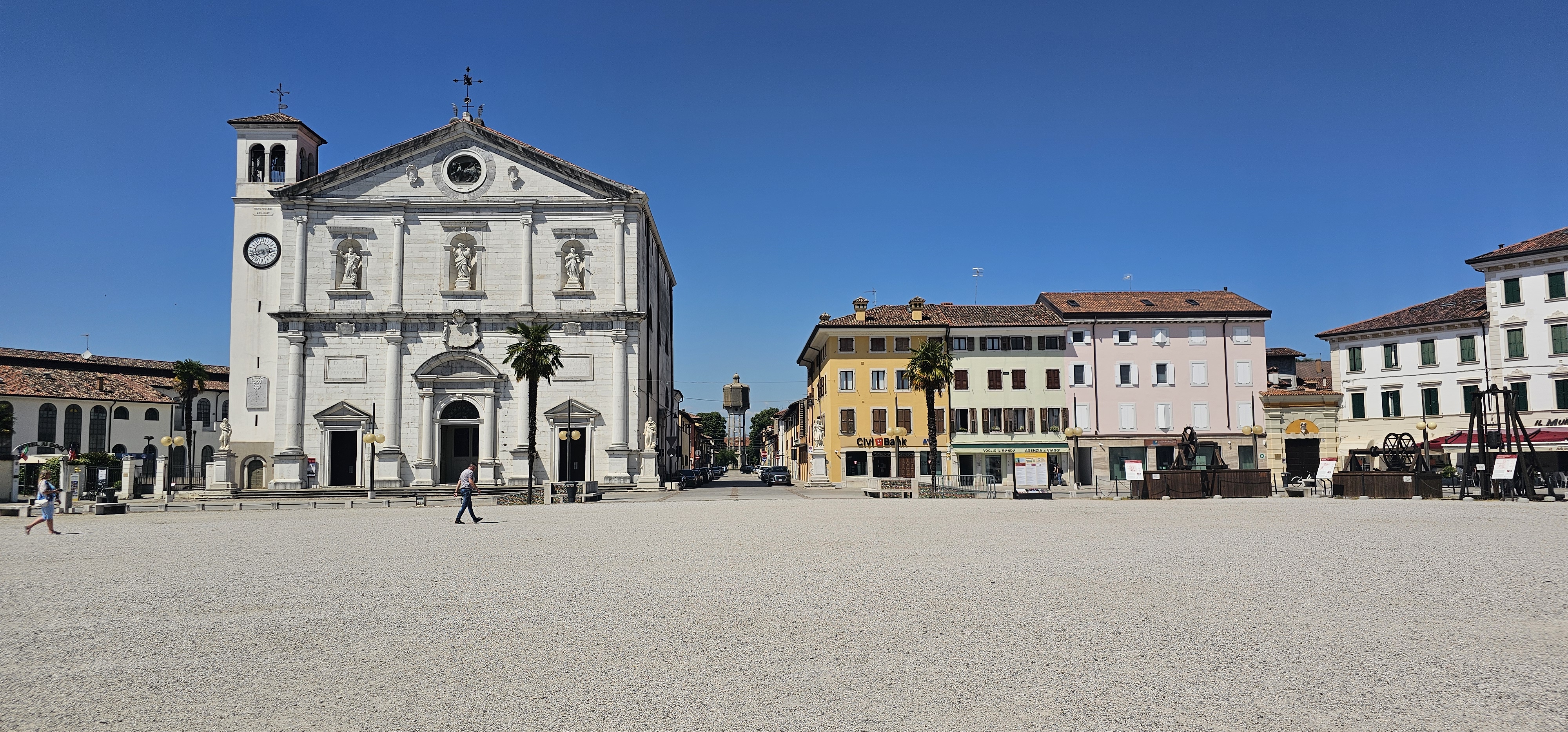 At the end of an open piazza, a white church with a stubby towns is next to a few shorter pastel builidings.